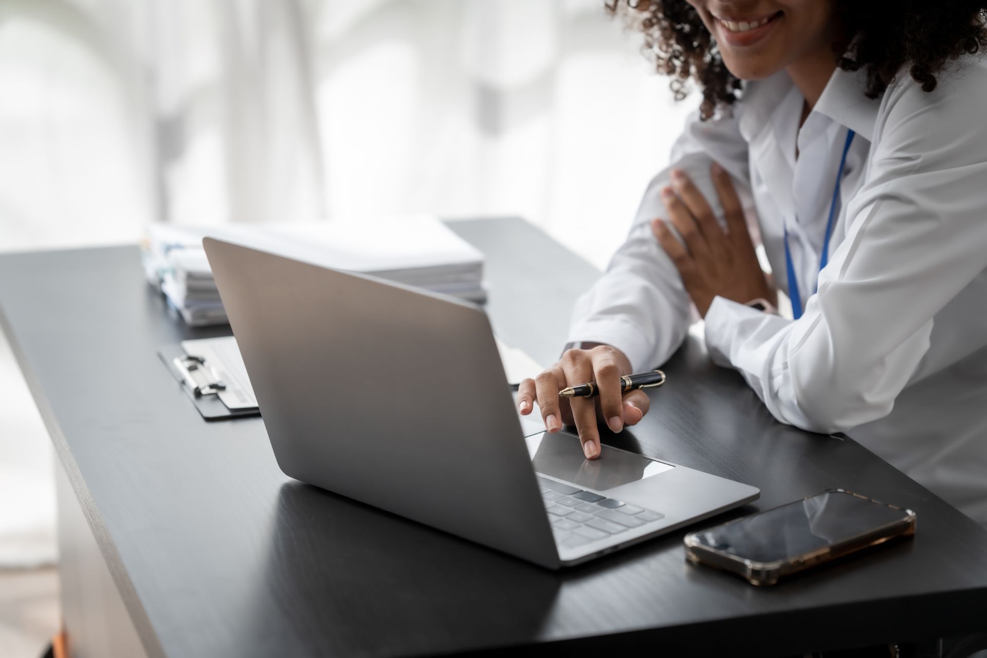 Doctor using laptop at a desk with phone and documents, smiling.