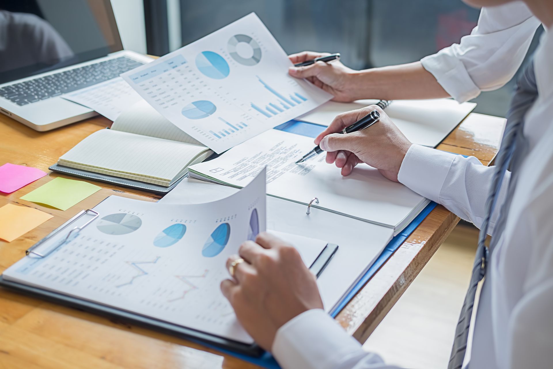 Two people reviewing charts and graphs at a desk with a laptop and open notebook.