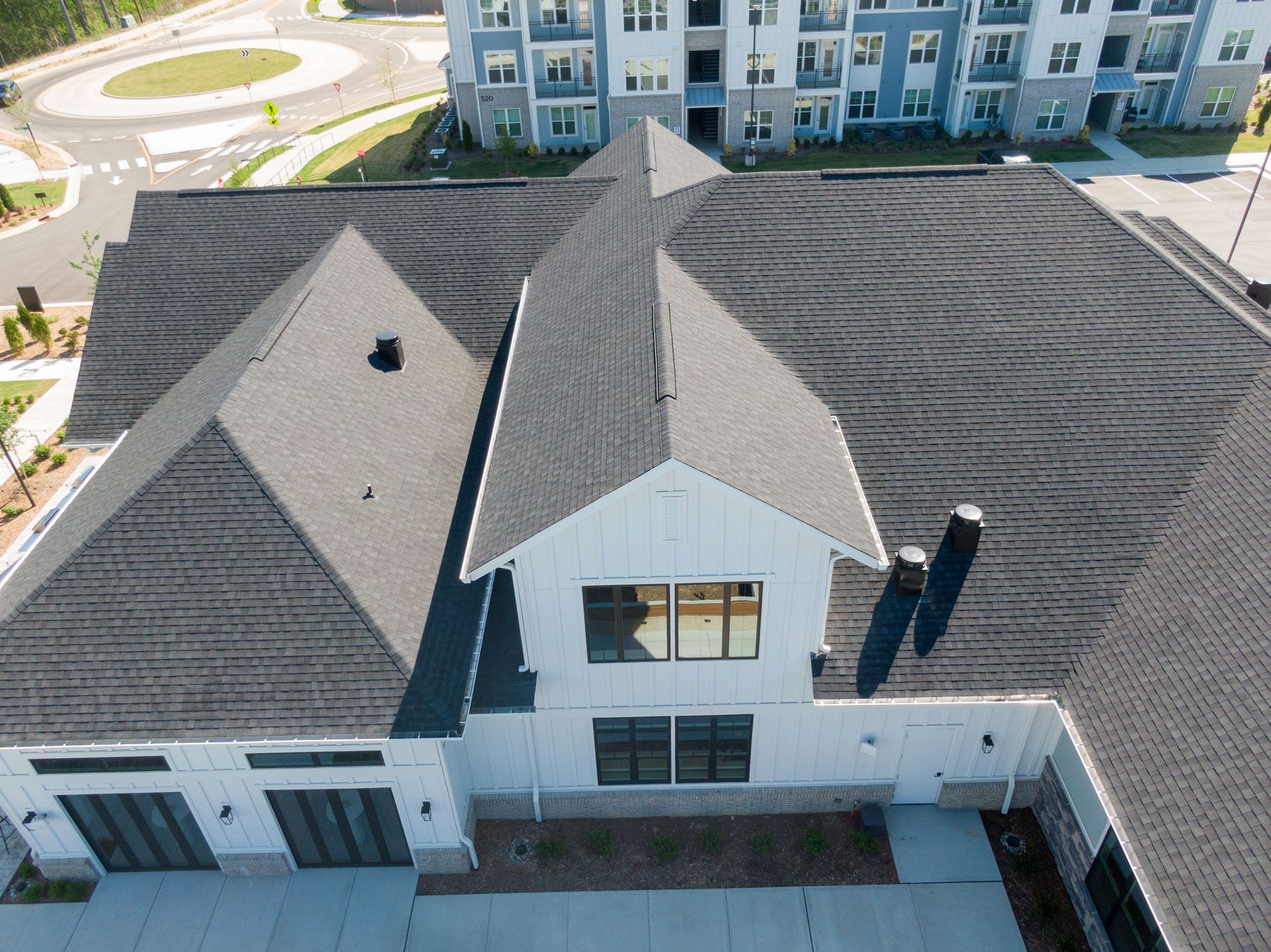 Aerial view of a white, modern apartment clubhouse with a complex gray shingled roof, set near a road and parking lot.