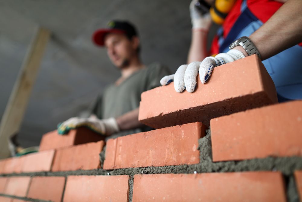 Bricklayer placing a red brick on a partly built wall; another worker in the background.