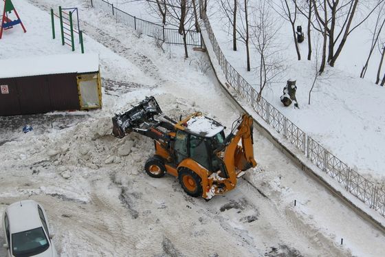 Yellow backhoe clearing snow from a residential area; snow on the ground, bare trees in the background.