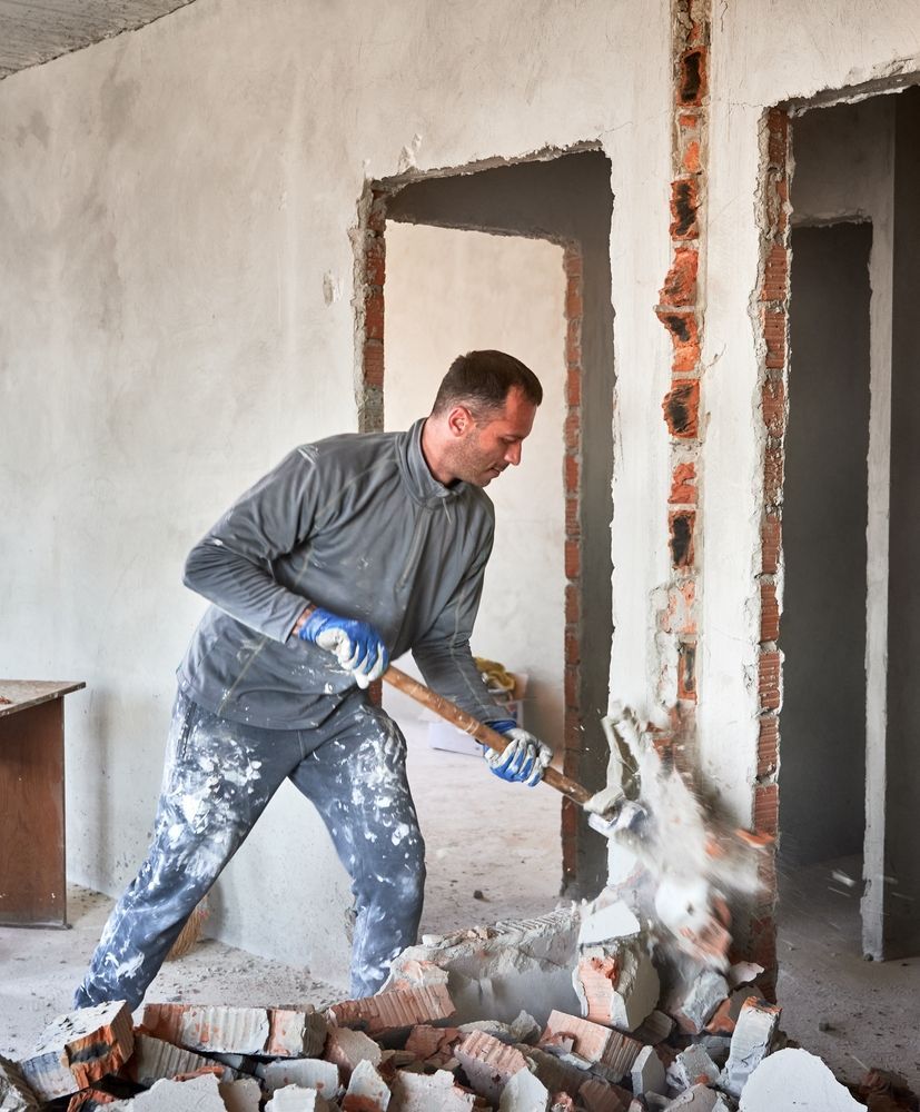 Man demolishing a brick wall with a tool inside a room, surrounded by rubble and dust. Man demolishing a brick wall with a tool inside a room, surrounded by rubble and dust.