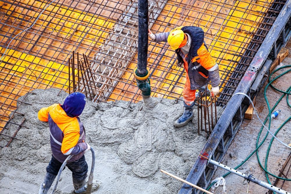Two construction workers pouring concrete from a hose, yellow and orange work attire, outdoor setting.