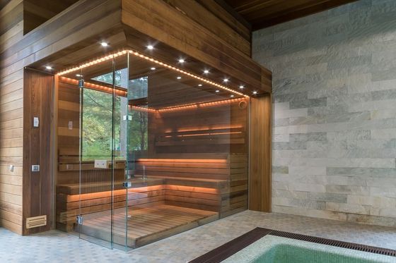 Modern wooden sauna with glass walls and warm lighting, adjacent to a pool.
