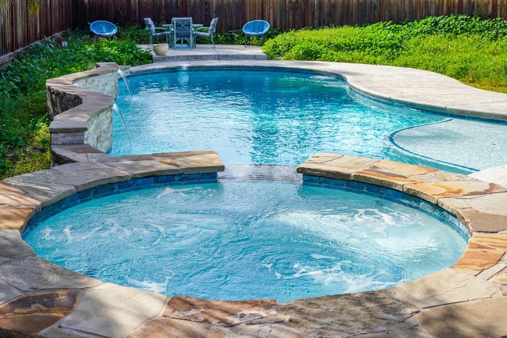 Swimming pool with attached jacuzzi surrounded by stone, with seating in background.