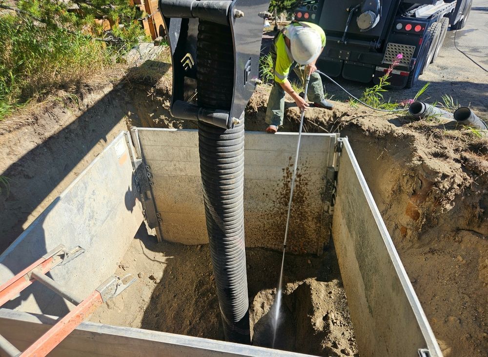 Construction worker in hard hat using a vacuum hose to clean a rectangular pit, with a truck in the background.