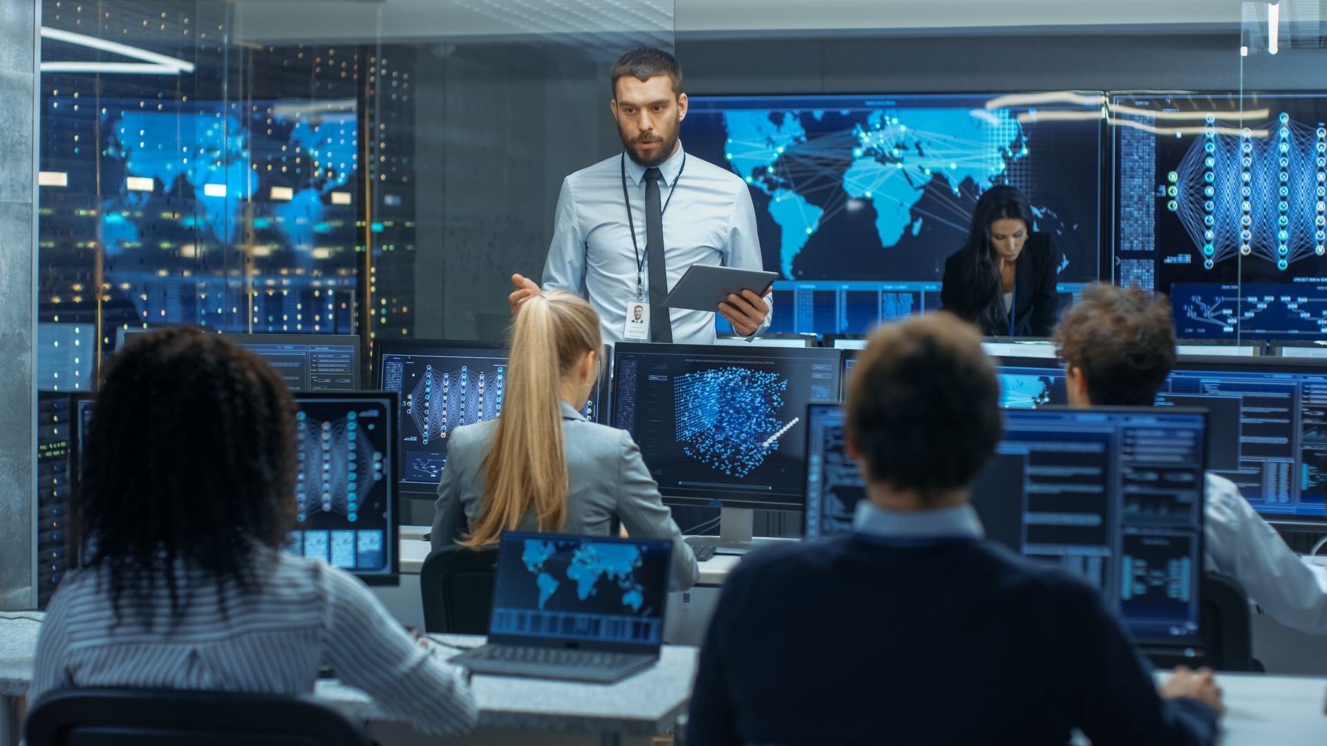 Man leading a team in a data center. Monitors show data and a world map. Team members are focused.