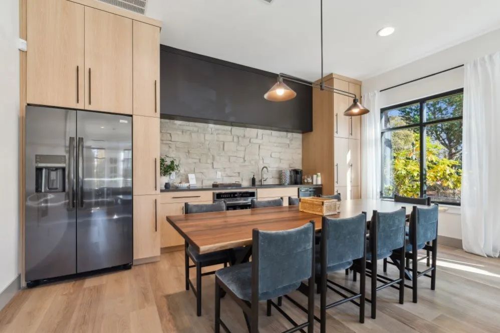 Modern kitchen with a wooden table and blue chairs, stainless steel fridge, light wood cabinets, and a stone accent wall at The Avery at Orenco Station, offers apartments near Orenco Station Town Center.
