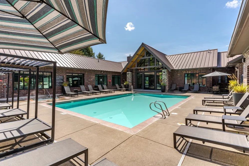 Pool area with lounge chairs, umbrella, and a building with a large glass window in the background at The Avery at Orenco Station, offers apartments near Orenco Station Town Center.