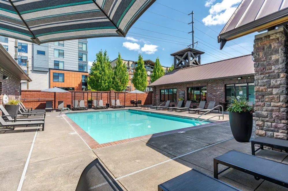 Swimming pool with lounge chairs under a striped umbrella at The Avery at Orenco Station, offers apartments near Orenco Station Town Center. Buildings and trees in the background.