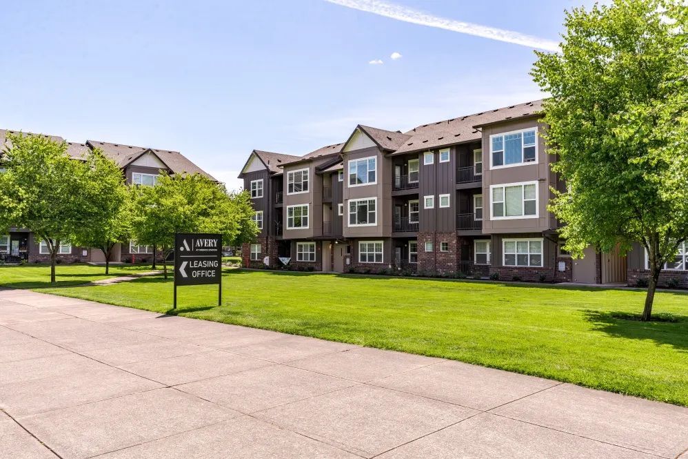 Apartment complex with brown siding, green lawn, sign, and trees on a sunny day at The Avery at Orenco Station, offers apartments near Orenco Station Town Center.