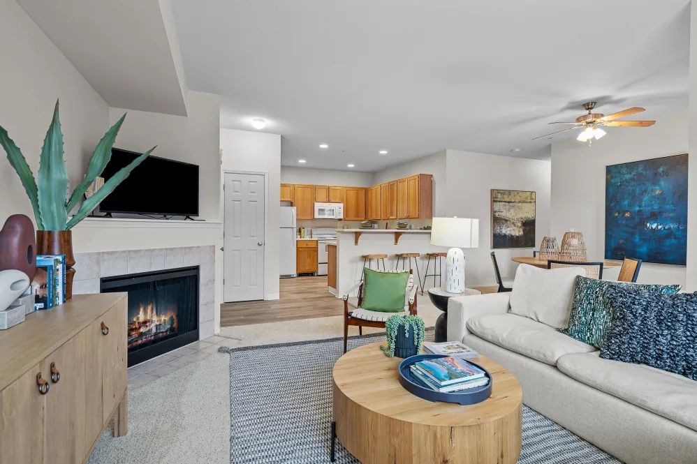 Living room with fireplace, open to kitchen at The Avery at Orenco Station, offers apartments in Hillsboro, OR. Beige sofa with blue pillows, wooden coffee table, TV over fireplace.