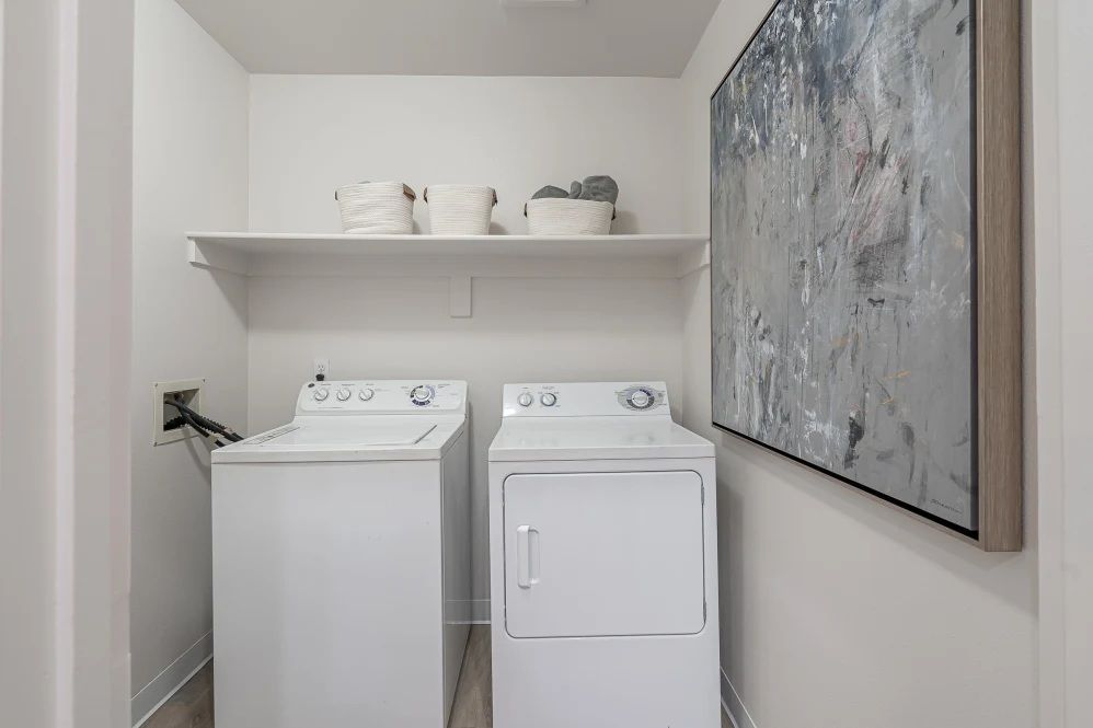 White laundry machines in a small, white-walled laundry room with a shelf, decor, and artwork at The Avery at Orenco Station, offers apartments in Hillsboro, OR. 