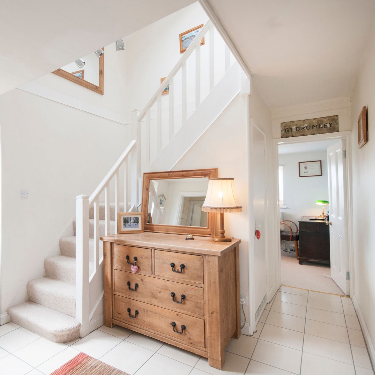 A bright white hallway with a staircase, wooden dresser, and open door to another room.