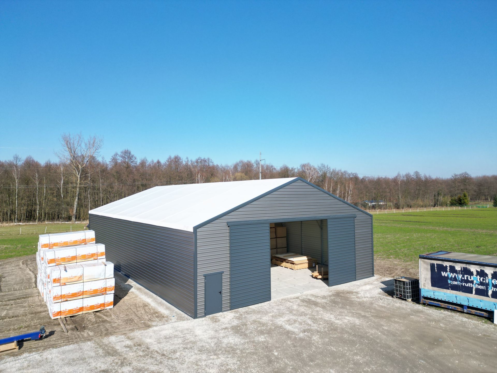 Szary metalowy magazyn z białym dachem na polu, w pobliżu stos materiałów budowlanych.