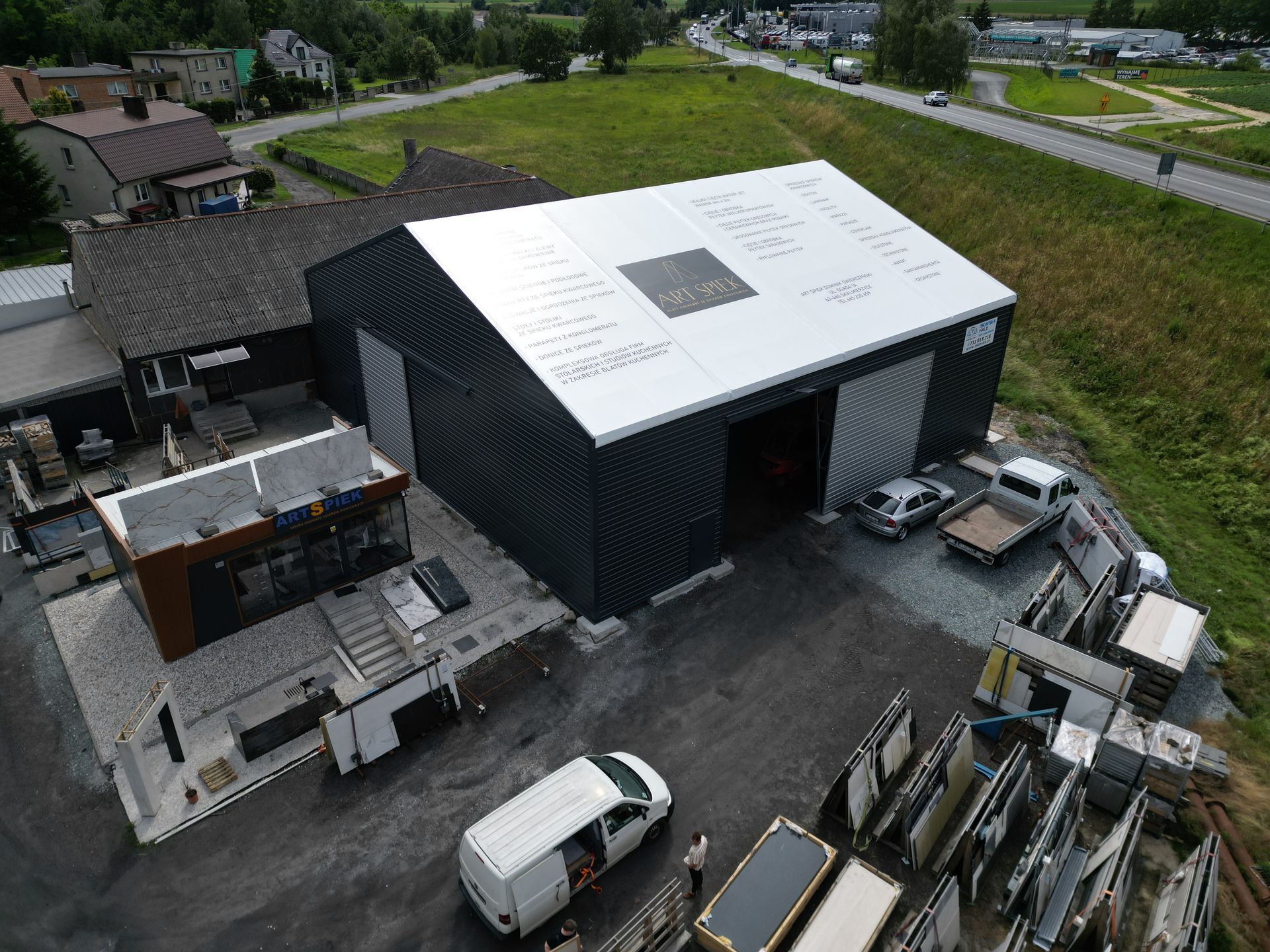 Widok z lotu ptaka na ciemny budynek przemysłowy z białym dachem, położony tuż przy parkingu i drodze.
