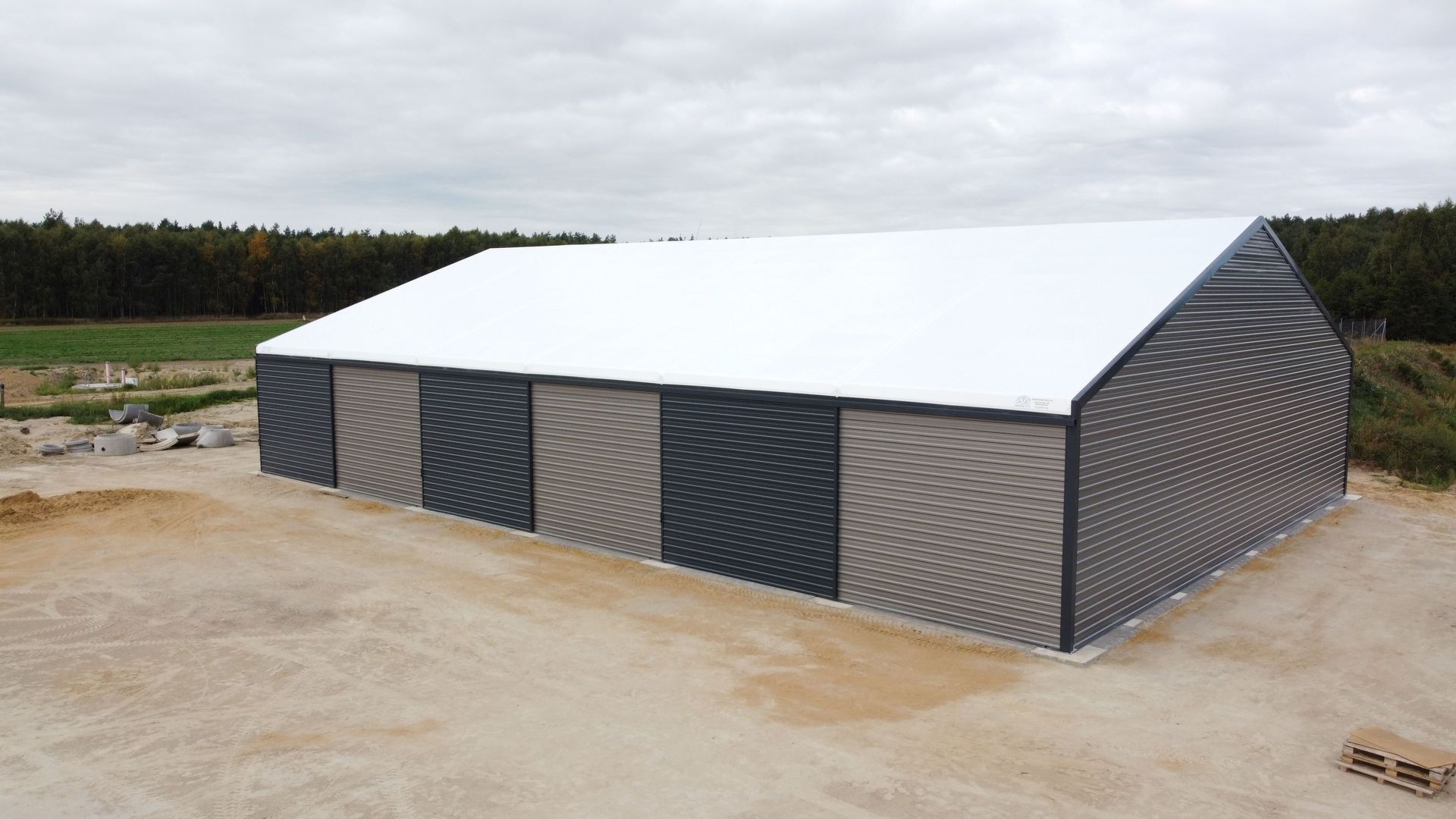 Duży, prostokątny budynek magazynowy z białym dachem i naprzemiennie szarymi i czarnymi ścianami z falistej blachy.
