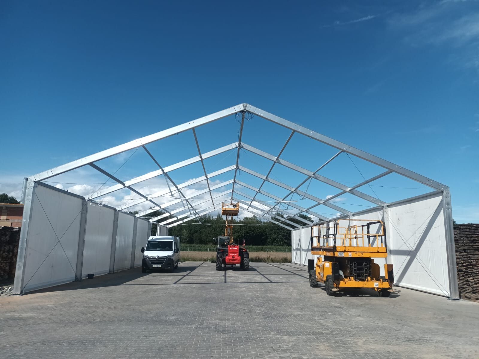 Metalowa konstrukcja z otwartymi bokami stoi na asfalcie, pod dachem znajduje się pojazd użytkowy i zaparkowany samochód dostawczy.