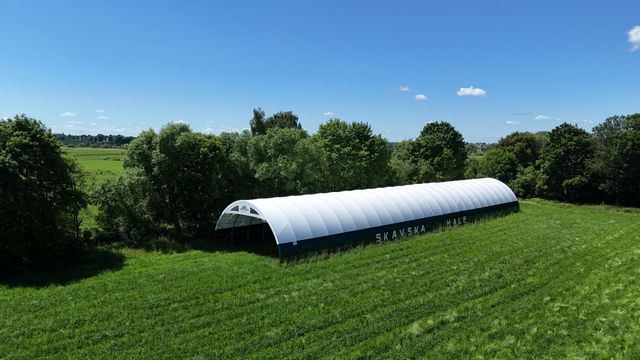 Długa, biała, łukowata konstrukcja z tkaniny stoi na zielonym polu otoczonym drzewami, pod czystym, błękitnym niebem.