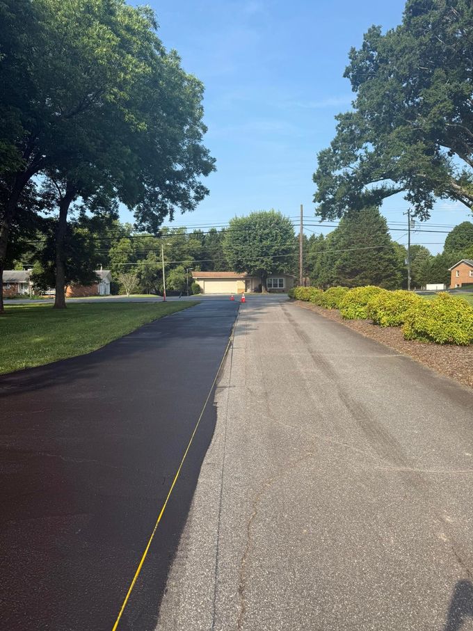 Asphalt driveway, newly paved on the left, old gravel on the right, bordered by grass and bushes, sunny day.