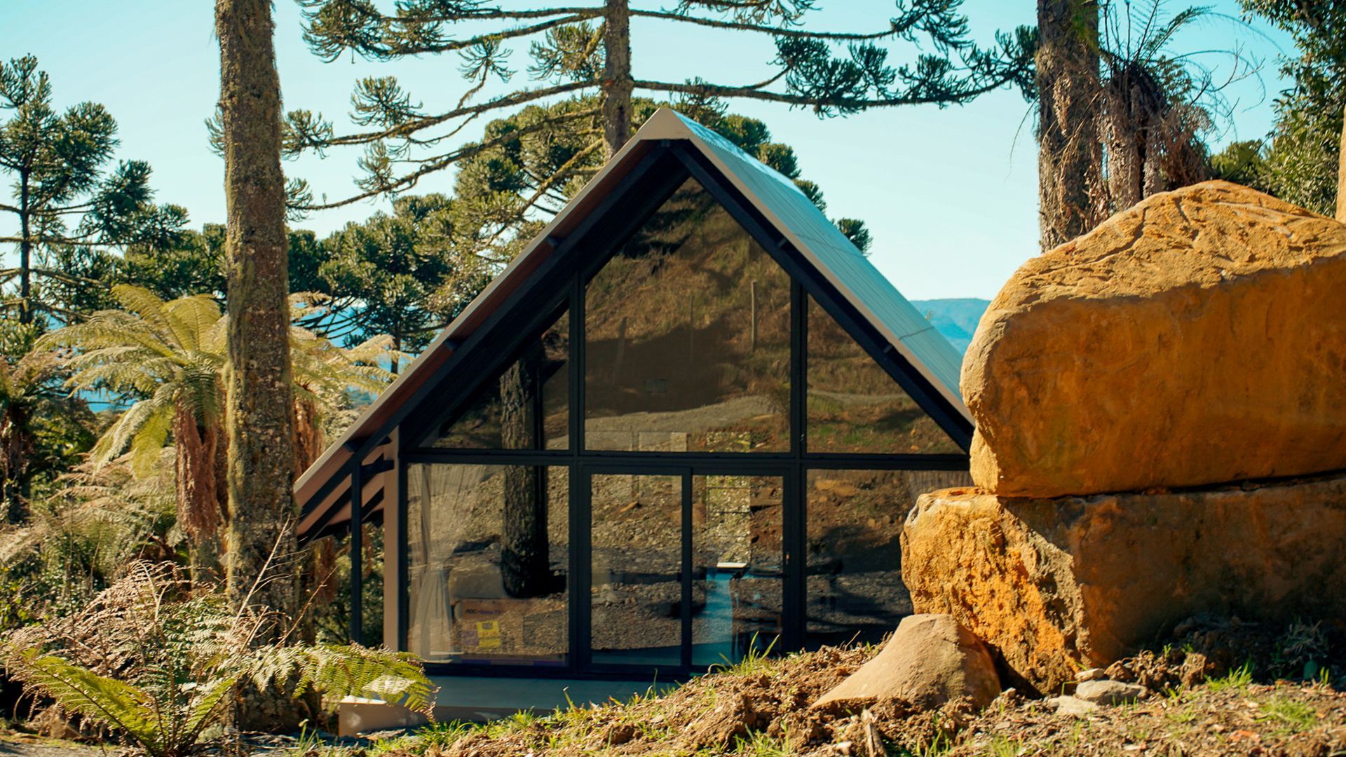 Cabana em formato de A com frente de vidro aninhada entre árvores e pedras em um dia ensolarado.
