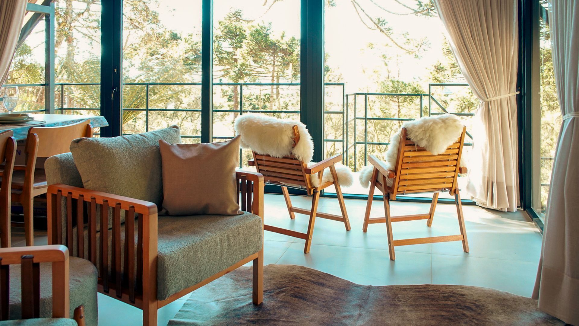 Sala de estar aconchegante com móveis de madeira e janelas do chão ao teto com vista para uma floresta. Duas poltronas com mantas de pele de carneiro.