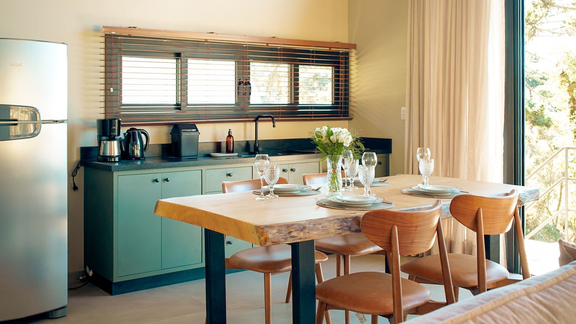 Uma cozinha rústica com uma mesa de jantar de madeira posta para uma refeição, ao lado de um balcão e uma geladeira. A paleta de cores é neutra, com toques de azul e madeira.