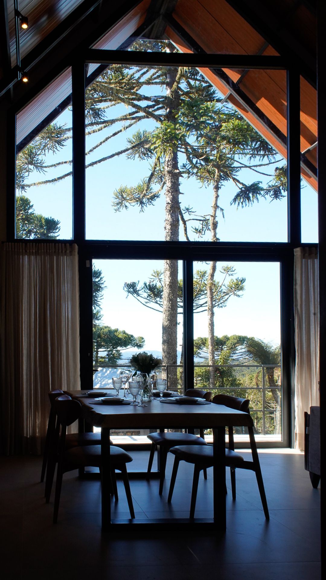 Espaço de jantar interno com grandes janelas emoldurando uma árvore alta contra um céu azul; mesa posta para jantar com cadeiras e cortinas.