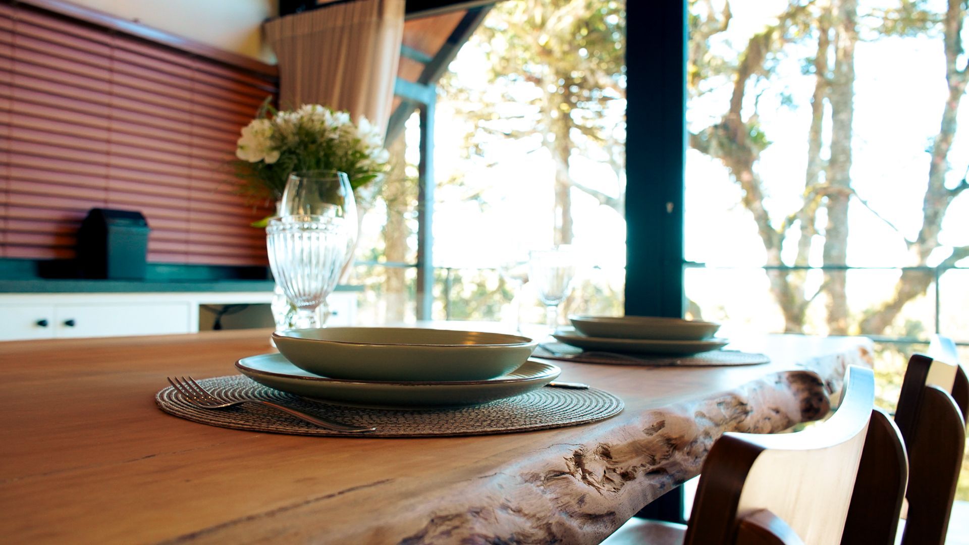Mesa de jantar posta com pratos e copos, ao lado de uma grande janela com vista para as árvores.