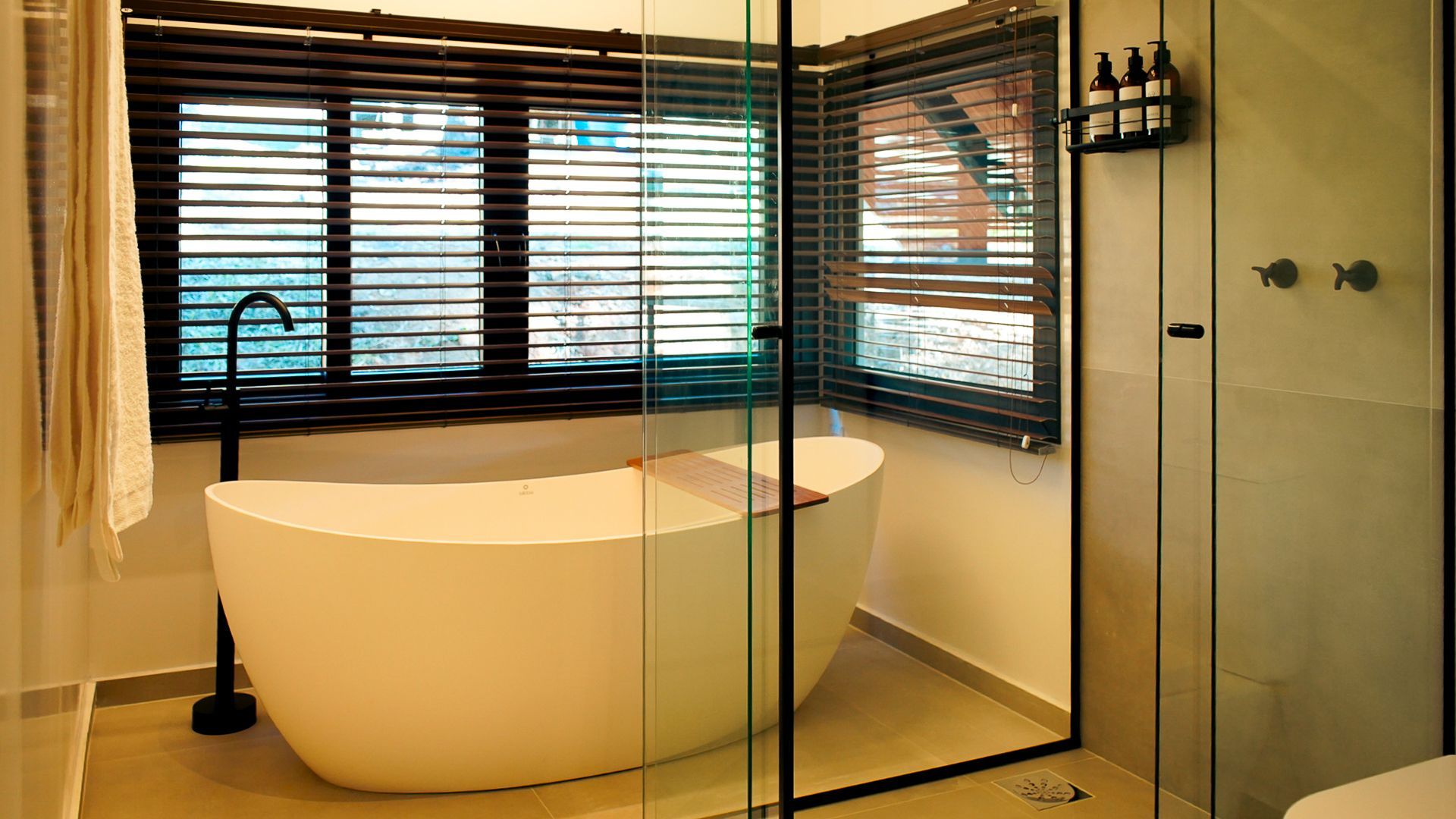 Banheiro moderno com banheira branca independente, box de vidro e persianas de madeira escura.