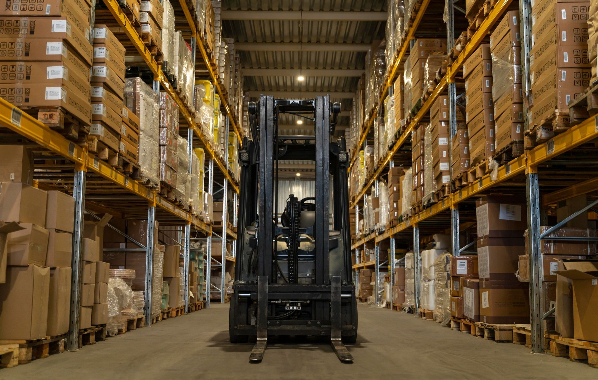 Forklift centered in a warehouse aisle between tall stacks of boxed pallets.