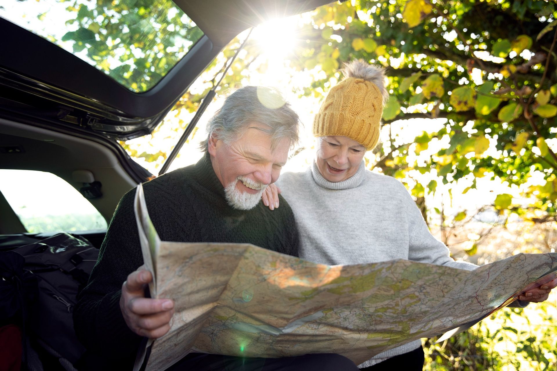 Two people study a folded map beside an open car trunk in a sunny wooded area