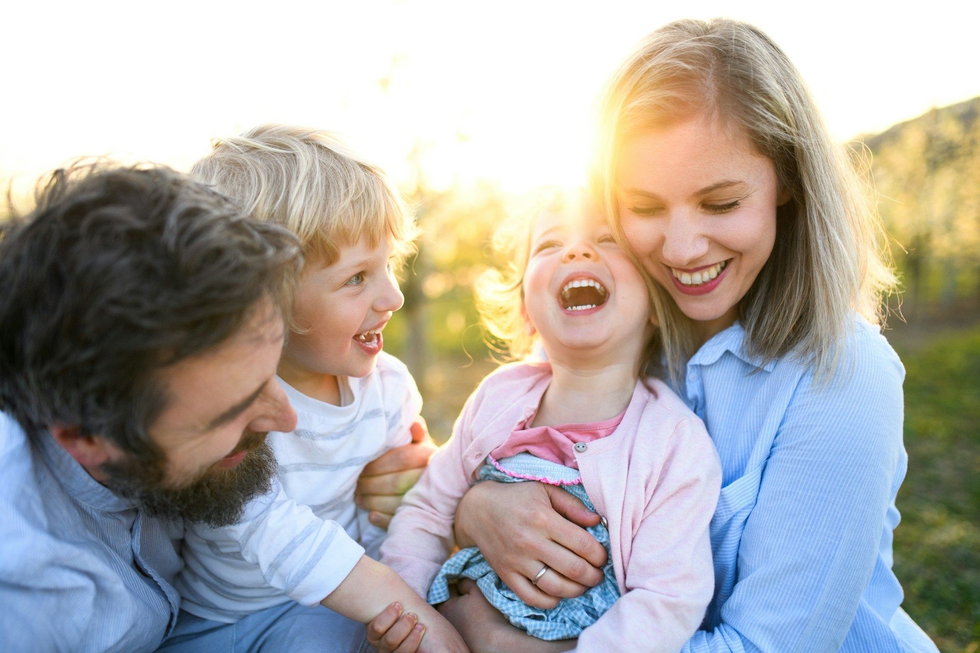 Family of four smiling outdoors in warm sunlight, with a young child laughing in the center