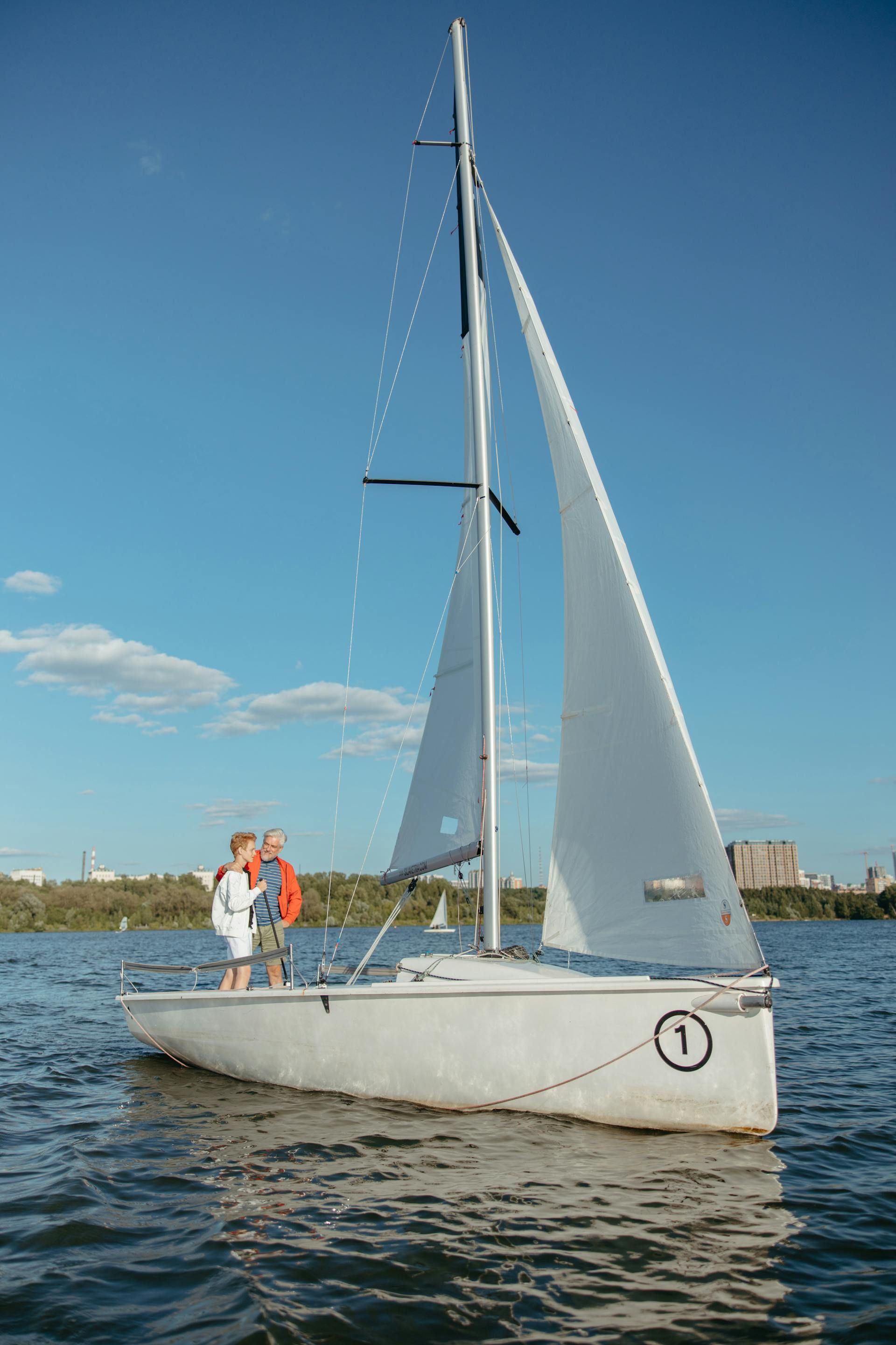 Two people standing on a white sailboat on calm blue water under a clear sky
