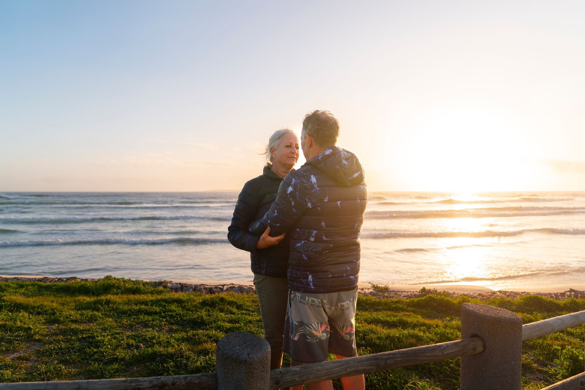 Two people standing by a seaside fence at sunset, facing the ocean in warm light