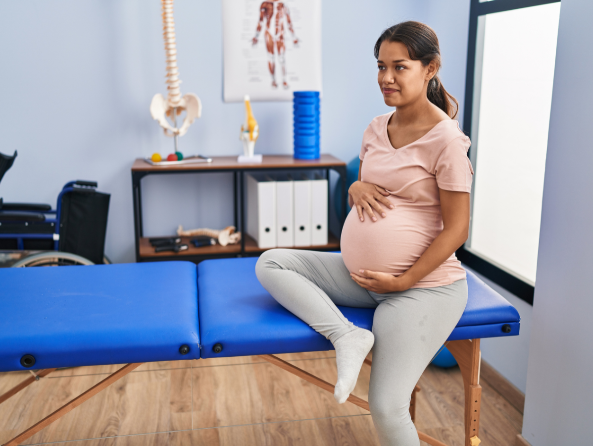 Pregnant woman sitting on a blue examination table, holding her belly in a medical office.
