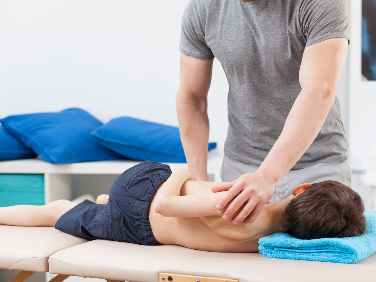Man assisting a child on a massage table; the child is lying down, possibly receiving physical therapy.