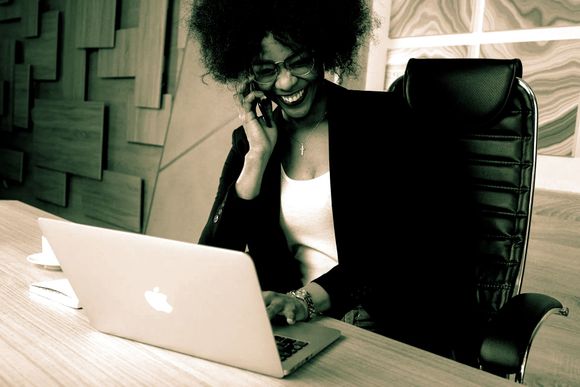 a woman is sitting at a desk using a laptop and talking on a cell phone .