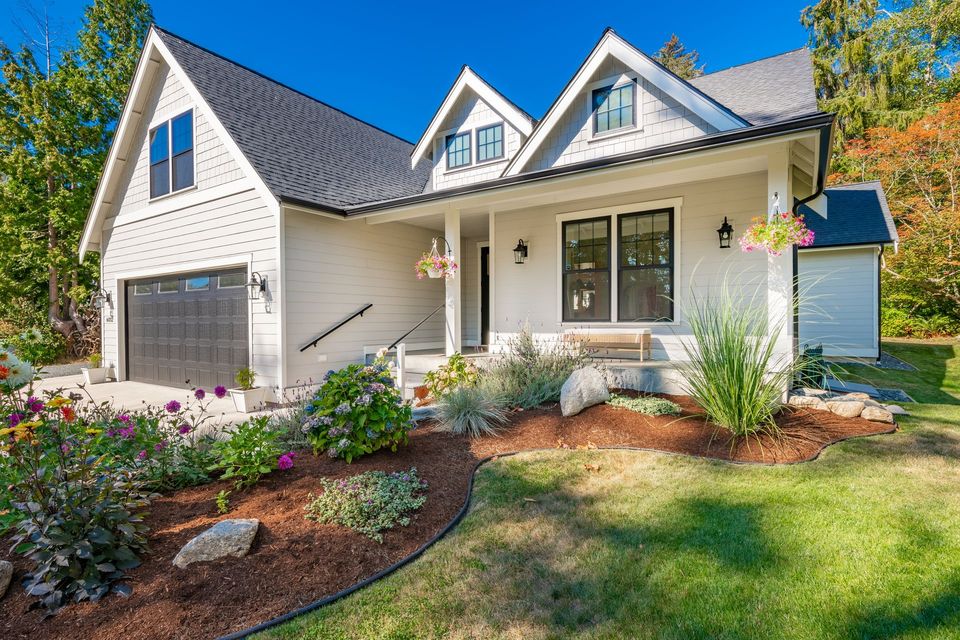 A white house with a black garage door and a porch surrounded by flowers.