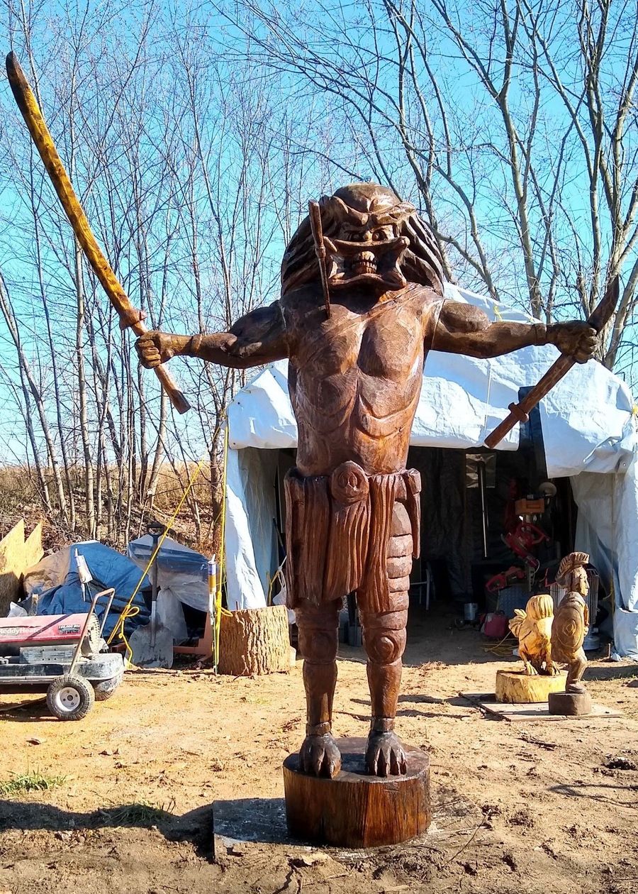Wooden statue of a bearded figure with outstretched arms in a sandy outdoor setting