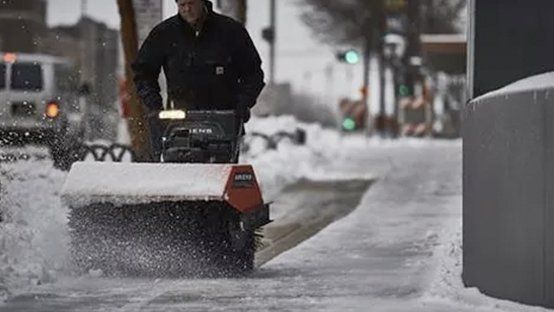 A man is pushing a snow blower down a snow covered sidewalk.