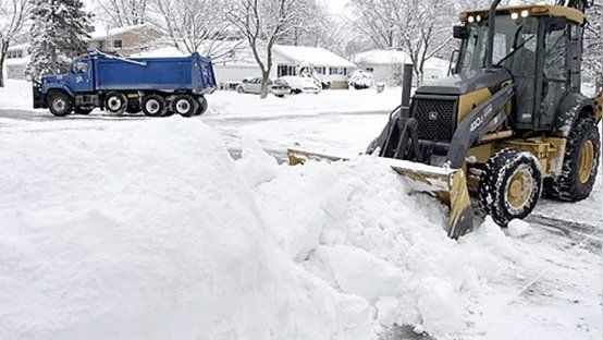 A snow plow is clearing snow from a driveway next to a dump truck.