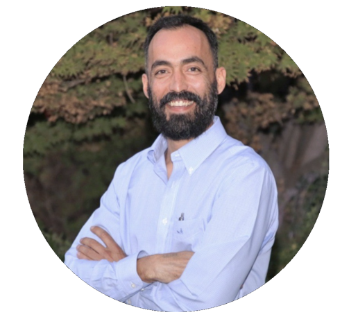 Man with beard, arms crossed, wearing light blue button-down shirt, smiling. Outdoors, circular frame.