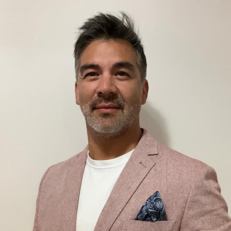 Hombre de cabello oscuro y expresión neutra, vestido con un blazer azul marino, sobre un fondo gris.