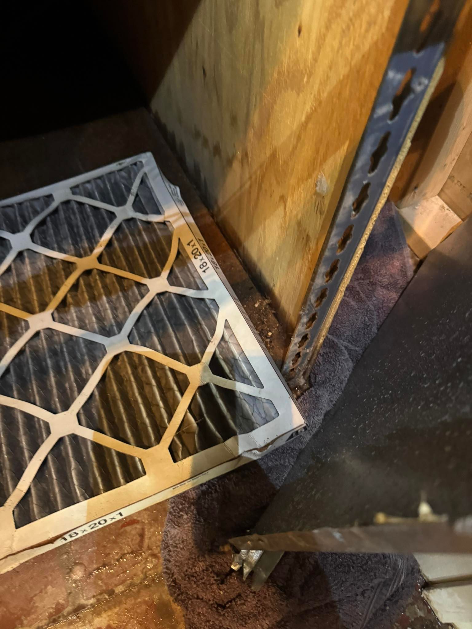 A pleated HVAC air filter rests on a floor next to a wooden wall and a metal support bracket.