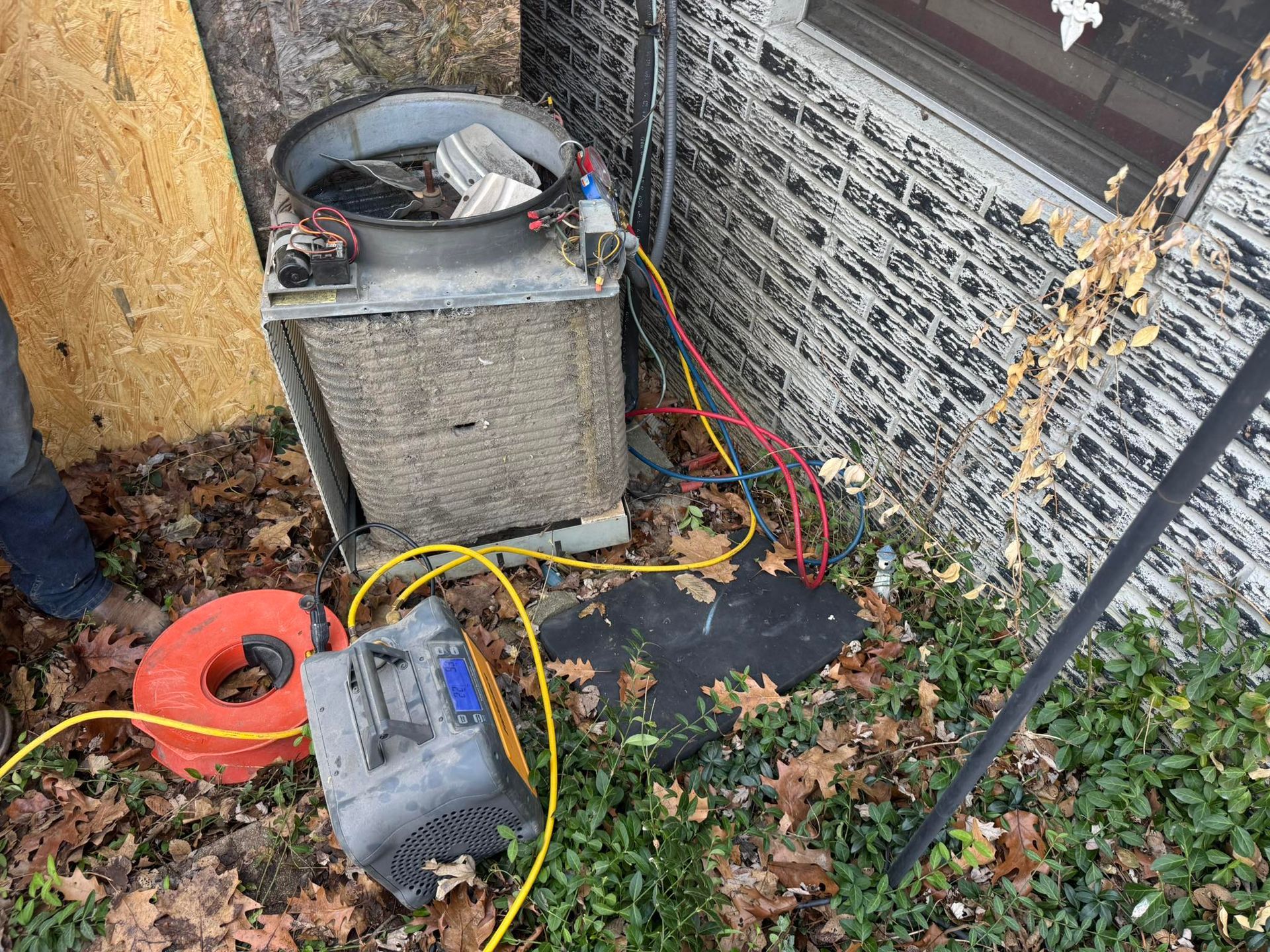 An HVAC technician services an outdoor air conditioning unit with gauges and hoses attached in a yard covered in leaves.