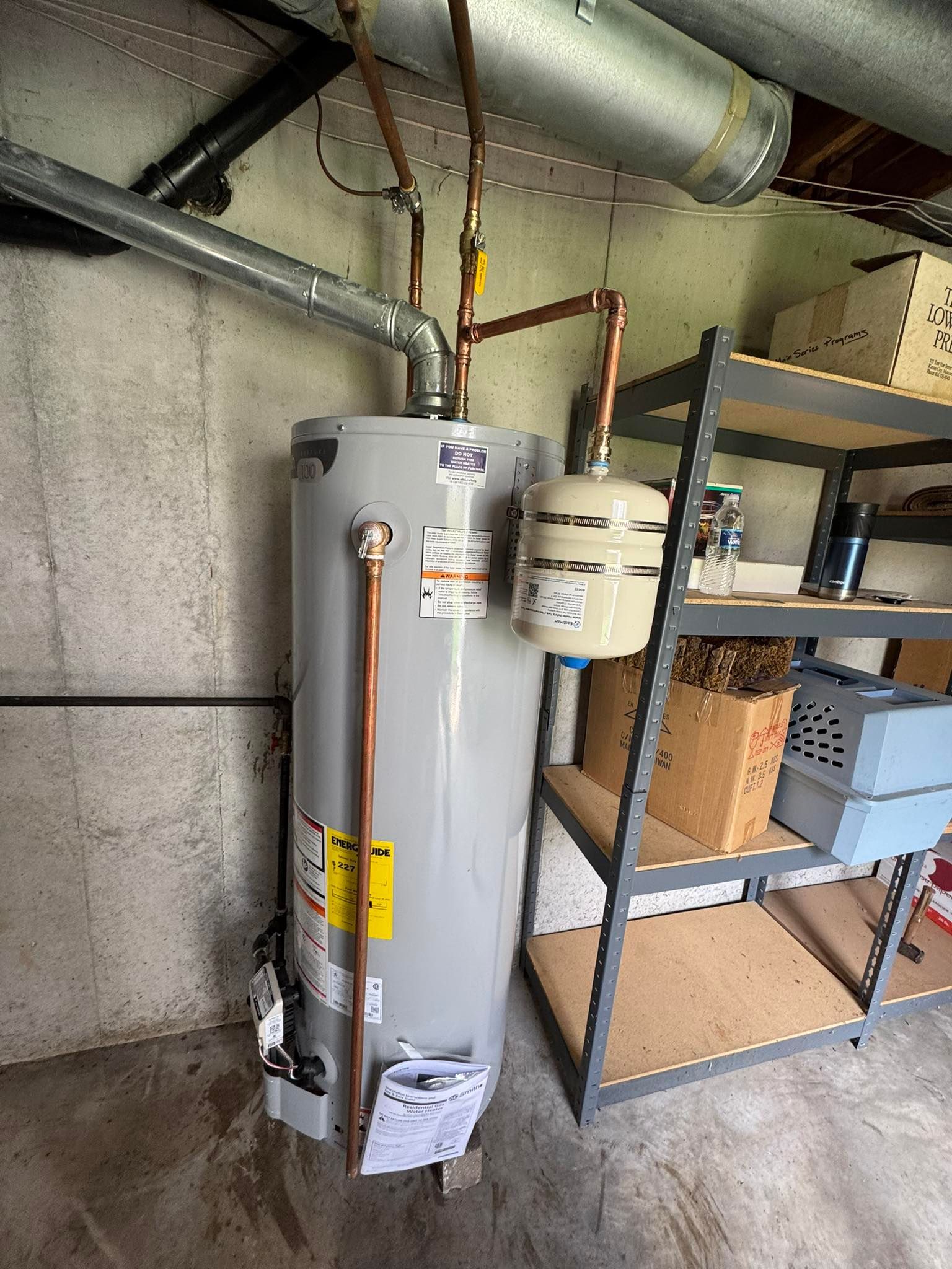 A gray water heater with a beige expansion tank installed in a basement next to metal shelving.