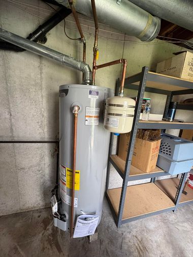 A gray water heater with a beige expansion tank installed in a basement next to metal shelving.
