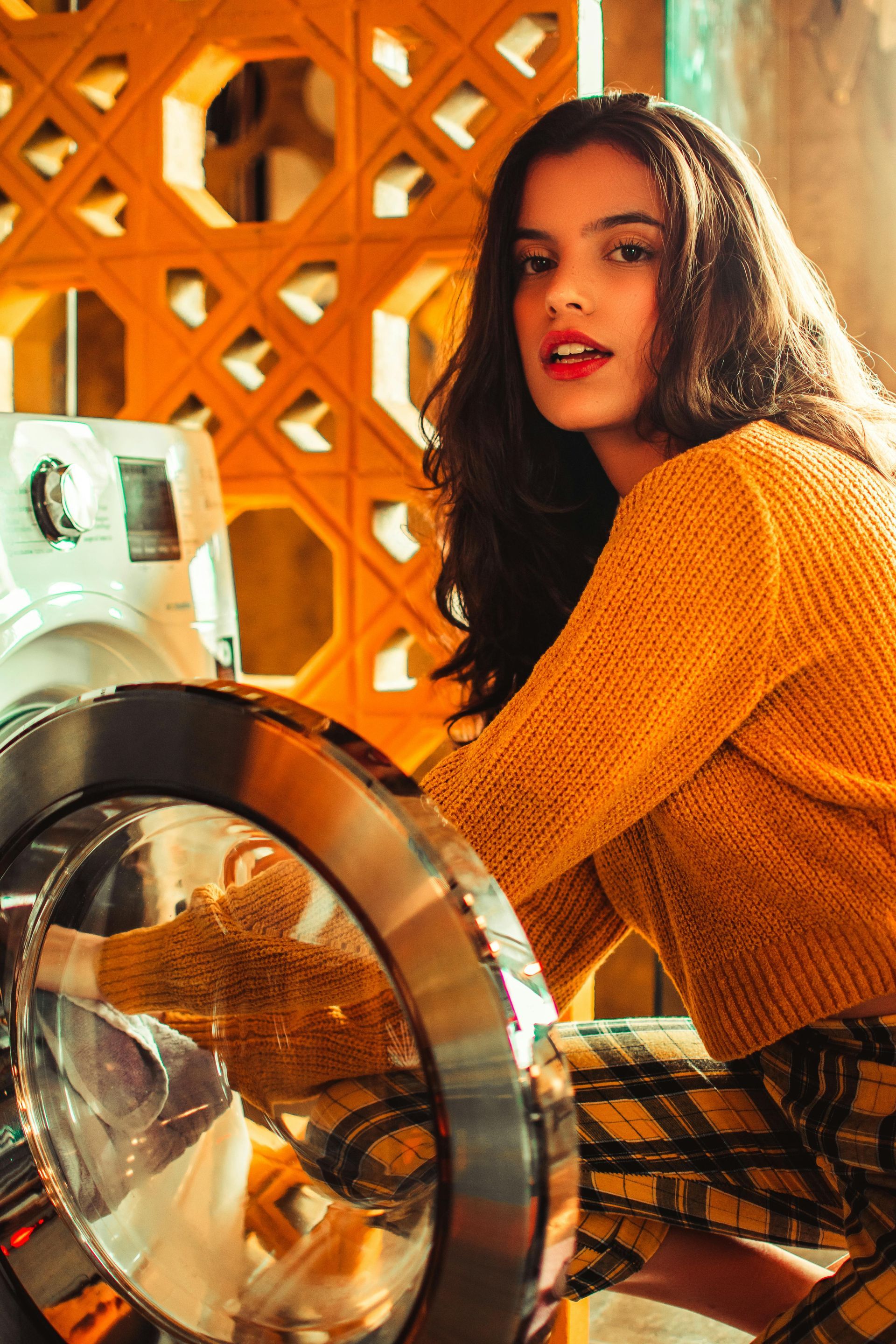 Woman in yellow sweater near washing machine, opening the door. Orange-toned setting.