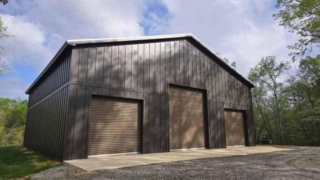 A large barn with three garage doors is sitting on top of a dirt road.